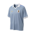 Camiseta retro de visitante de Francia 1982 de Hinchas Sportswear Store, de color azul grisáceo con cuello blanco, ribetes en las mangas y detalles de rayas en los hombros.